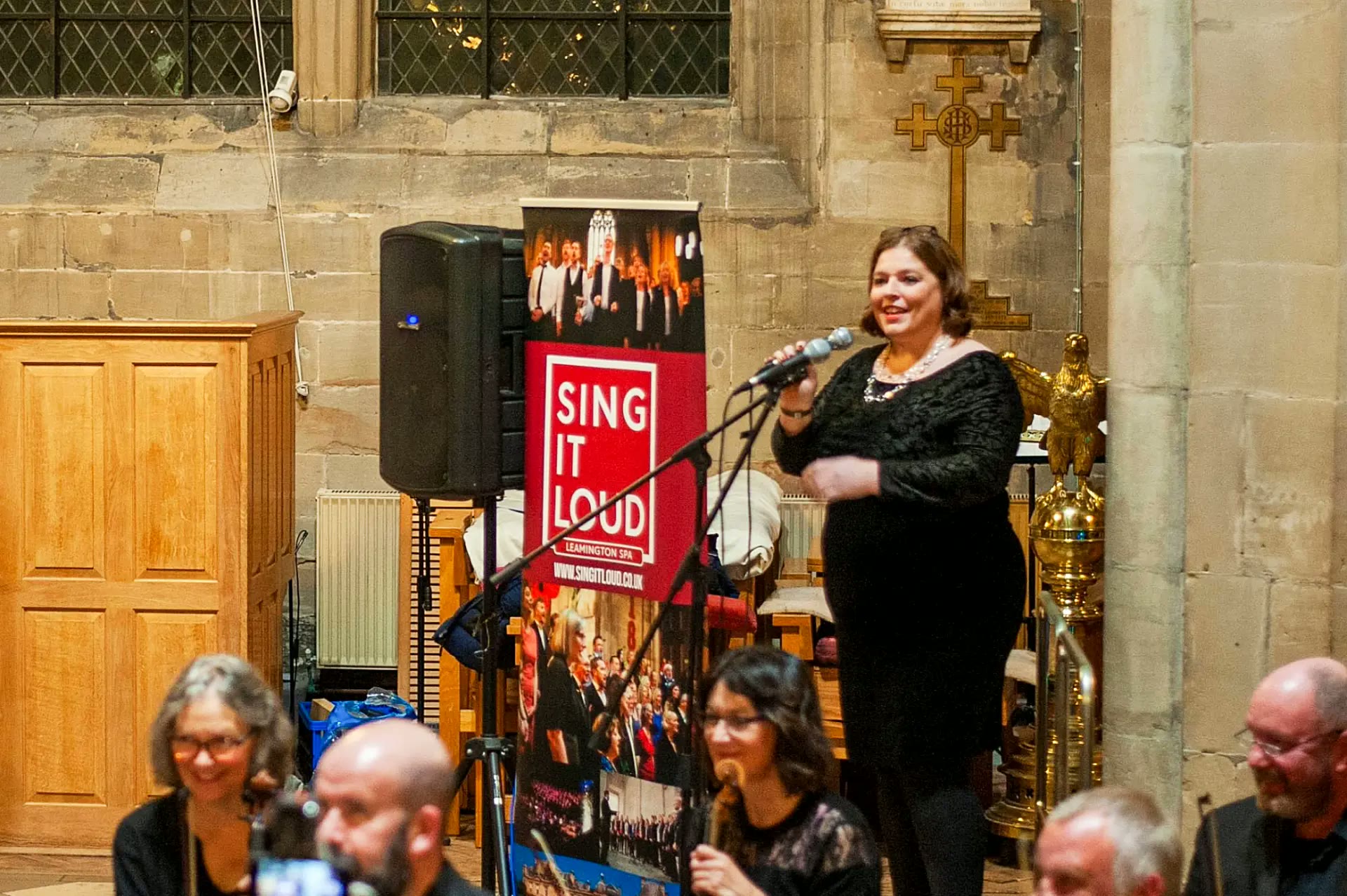 Rosie Sayers speaking warmly into a microphone at a church event, with a Sing It Loud choir banner behind her